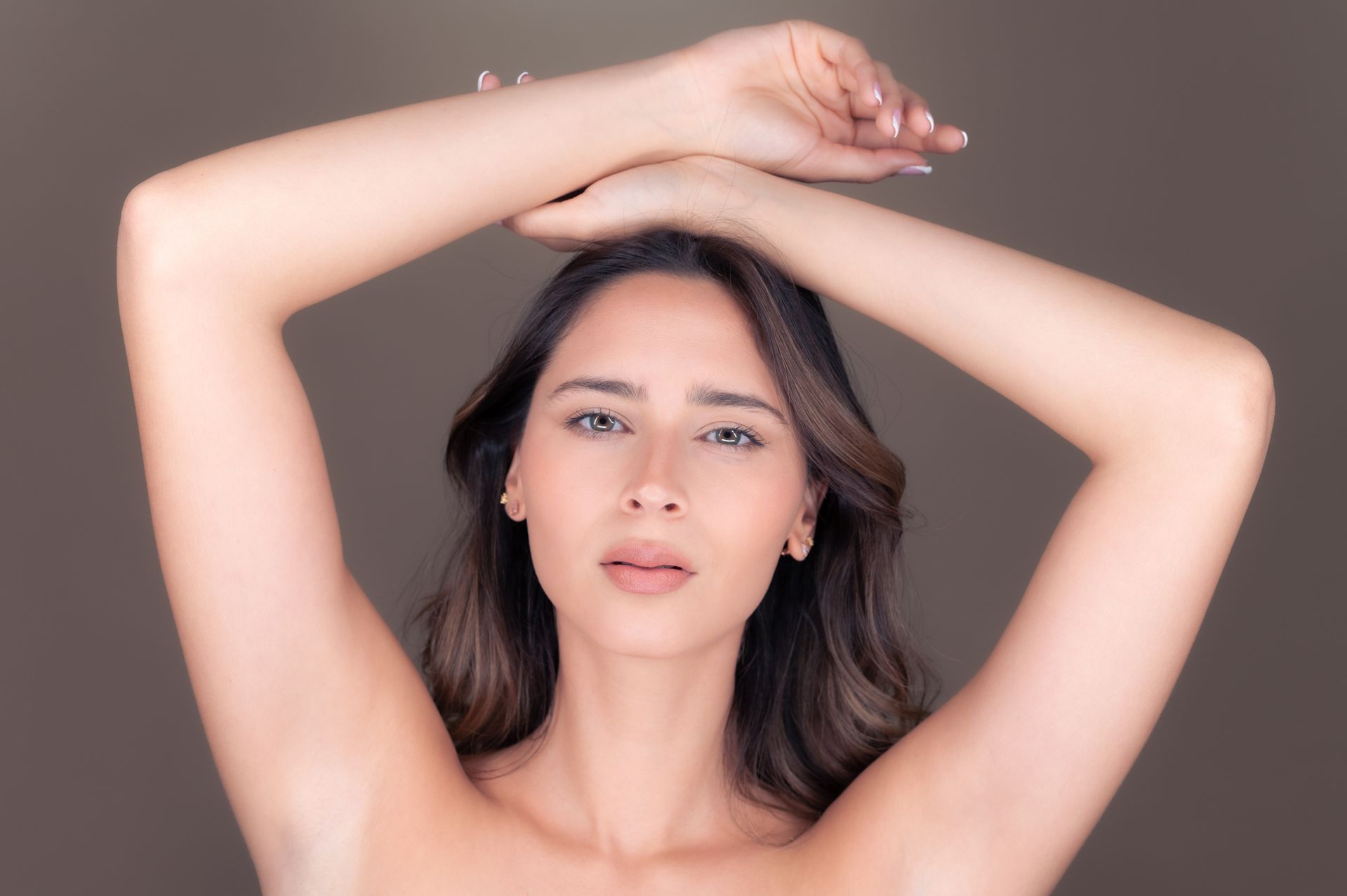 A woman with long brown hair raises her arms above her head, crossing her wrists. She has a neutral expression and appears against a plain, soft brown background.